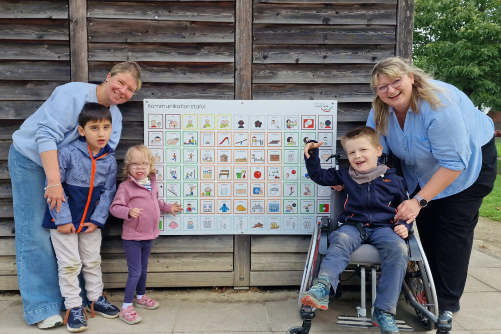 Veronika Bense (r.) und Sabine Leson präsentie-ren mit einigen Kita-Kindern die neue Kommunikationstafel. Foto: Stiftung Haus Hall